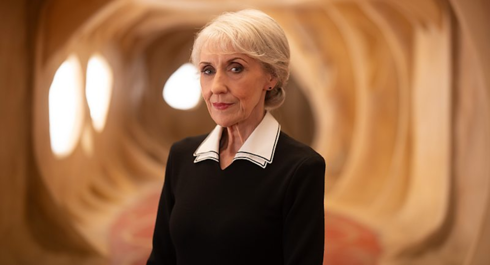 Anita Dobson as Mrs. Flood stands in a well lit beige hallway